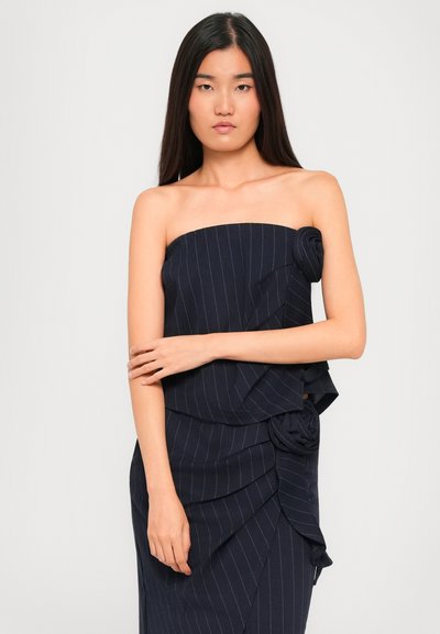 Jonge vrouw met langdurig donker haar, gekleed in een strapless marineblauwe gestreepte top en bijpassende rok met stoffen bloemen details, staand tegen een witte achtergrond.