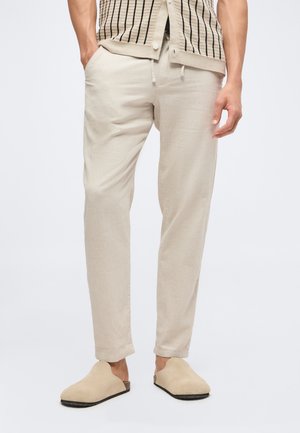 Homme portant un pantalon en lin beige clair avec des poches latérales, un gilet en maille rayé et des chaussures beige assorties sans lacets, debout devant un fond uni.