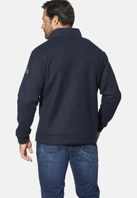 Marineblaue Fleecejacke mit hohem Kragen, strukturierter Stoff, gepolsterten Schulterakzenten und einem dezenten Logopatch am linken Ärmel.