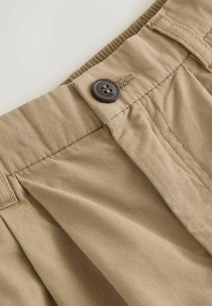 Nærbillede af beige bomuldsbukser taljebånd med syet knap og bæltestropper, der viser stoftekstur og skræddersyede sømme.