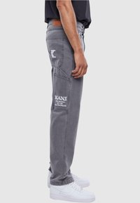 Pantalones de mezclilla gris con bolsillos laterales, marca bordada en blanco y costuras distintivas. Llevados con zapatillas blancas.