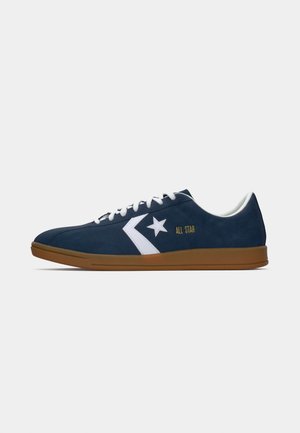 Tamno plave suede tenisice s bijelim detaljima, logotipom zvijezde i oznakom "ALL STAR"; imaju smeđu gumenu potplat i bijele vezice.