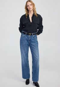 Camicia nera con bottoni e due tasche sul petto, abbinata a jeans blu a gamba dritta a vita alta. Cintura nera con una fibbia dorata rettangolare.