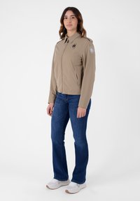 Giacca leggera beige con chiusura a zip, due tasche laterali e un patch con logo sulla manica. Abbinata a jeans blu e sneakers bianche.