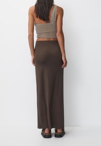 Een vrouw staat met de rug naar ons toe, gekleed in een taupekleurige crop top met één schouder, een lange donkerbruine rok en bruine instapschoenen met studs.