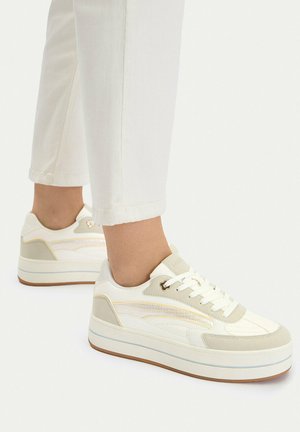 Osoba nosząca białe sneakersy na platformie z beżowo-złotymi akcentami oraz białe krótkie spodnie na jednolitym jasnym tle.