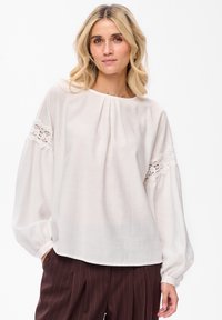 Kobieta z blond włosami, ubrana w białą bluzkę z koronkowymi detalami na rękawach oraz ciemne spodnie w prążki, stojąca przed białym tłem.