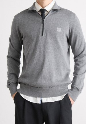 Man wearing gray quarter-zip sweater over white shirt and black tie, with black pants, standing with hands in pockets.