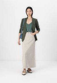 Olijfgroene blazer, geribbelde grijze maxirok en beige schoenen. Model draagt een kleine blauwe tas. Eenvoudige, aansluitende vorm met een golvende halslijn.