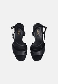 Sandali in pelle nera con un design a strappi, dotati di un leggero tacco, punta aperta e dettaglio del logo dorato sulla suola. Fibbia regolabile sulla cinghia.
