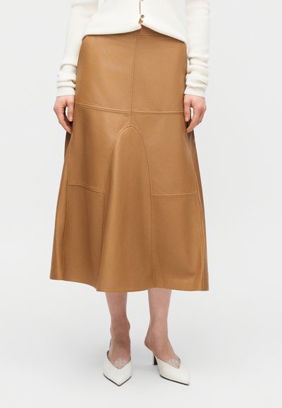 Γυναίκα που φοράει μια μπεζ δερμάτινη μίντι φούστα με ραφές πάνελ, λευκό μακρυμάνικο τοπ και λευκές μυτερές γόβες με λεπτομέρεια δαντέλας.