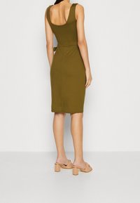 Robe ajustée vert olive avec un dos en décolleté arrondi, un détail ceinturé à la taille, et une longueur sous le genou. Le mannequin porte des mules à talons blocs beige.