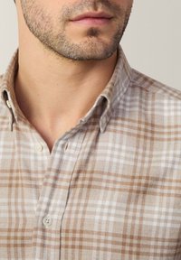 Camisa de cuadros en tonos beige y marrón, hecha de una tela suave. Cuenta con un cuello americano y botones blancos, con una textura acogedora y un corte entallado.