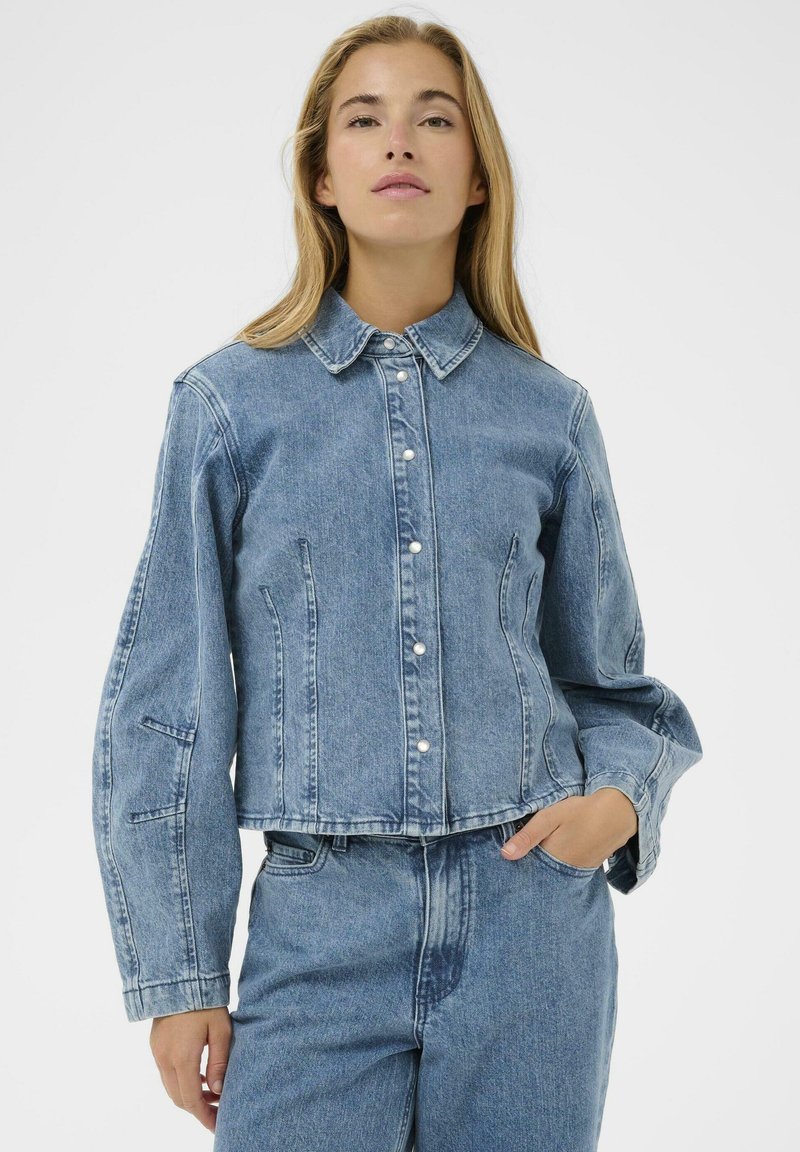 Femme aux cheveux blonds longs portant une veste en denim bleue boutonnée avec des boutons pression en perle et un jean taille haute assorti, une main dans la poche.