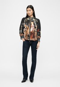 Femme portant une veste noire avec une impression détaillée de style Renaissance, un jean foncé et des talons noirs pointus, debout les mains dans les poches.