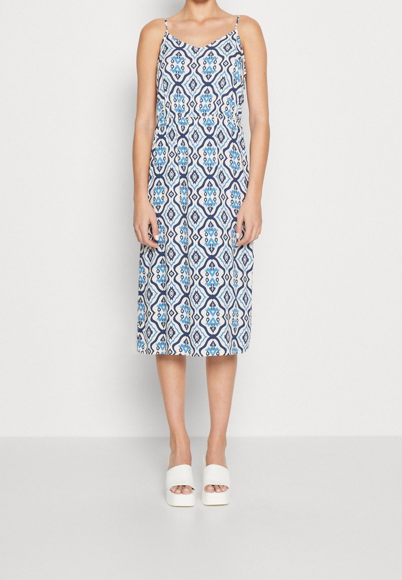 Femme portant une robe sans manches, longueur genou, avec des motifs géométriques bleus et blancs et des chaussures compensées blanches à bout ouvert.