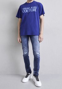 Camiseta azul de gran tamaño con el texto "VERSACE JEANS COUTURE", combinada con jeans de mezclilla rasgados y zapatillas blancas con cordones negros.