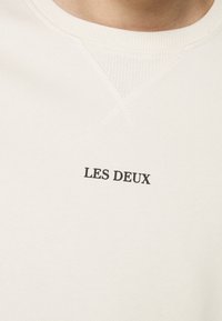 Light beige sweatshirt with ribbed neckline. "LES DEUX" printed in black on the chest. Soft texture, casual design, minimalistic style.