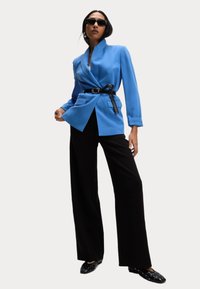 Blauwe op maat gemaakte blazer met een ceintuur in de taille, gecombineerd met zwarte wijde broekspijpen en versierde zwarte schoenen, tegen een neutrale achtergrond.