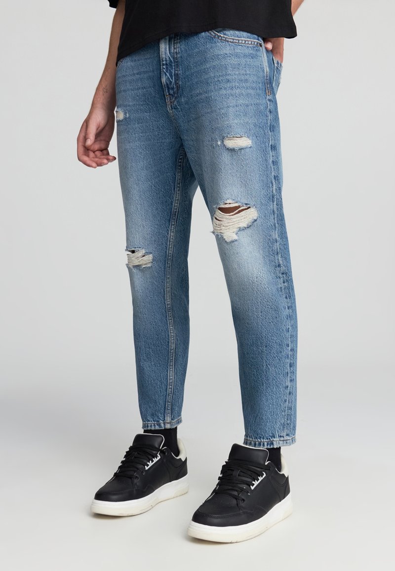 Jeans in denim chiaro, strappati, con una vestibilità comoda, caratterizzati da orli sfilacciati e dettagli distrutti. Abbinati a sneakers nere.