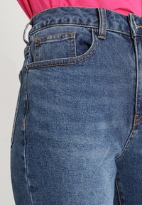 Jeans de tiro alto, de color azul, hechos de mezclilla con costuras visibles, un bolsillo delantero y una textura suave. El color muestra áreas desgastadas.