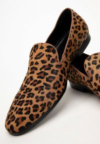 Světlé slip-on boty s leopardím potiskem, hnědým a béžovým texturovaným povrchem, s hladkou černou podrážkou a zaoblenou špičkou.