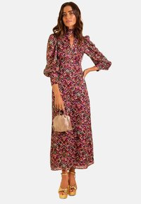 Vestido maxi con un colorido patrón ikat, mangas largas con voluminosidad, cuello alto y detalles plisados. Combinado con un bolso rosa suave y tacones de plataforma dorados.