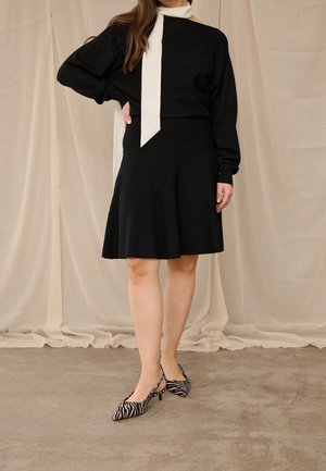 Femme portant une robe noire à manches longues avec une écharpe blanche nouée au cou, debout avec une main sur la hanche, talons imprimé zèbre sur un sol en béton.