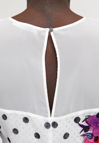 Blouse blanche avec une fermeture en goutte d'eau au dos, motif à pois et accents floraux à l'ourlet. Dotée d'un tissu texturé.