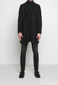 Svart överrock i ullblandning med skräddarsydd design, två knappar och en enkel bröstficka, kombinerad med passande svarta byxor och skor.