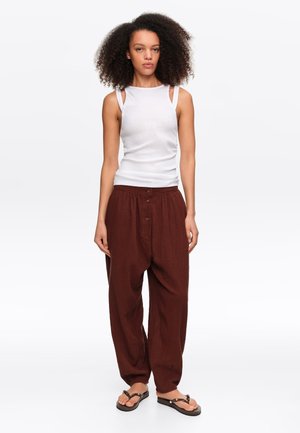 Kobieta z kręconymi włosami, ubrana w biały top bez rękawów, luźne brązowe spodnie z guzikami i czarne klapki, stojąca na białym tle.