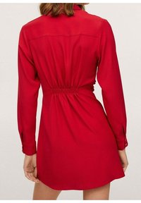 Robe rouge à manches longues, taille élastique et poignets boutonnés. Tissu lisse, coupe au genou et détail de empiècement au dos.