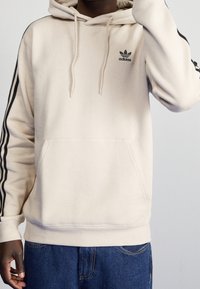 Beige hoodie av mjukt tyg med en känguruficka, dragsnören och svarta ränder längs ärmarna. Har en svart logga på bröstet.