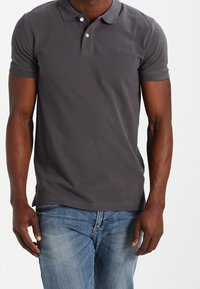 Man som bär en mörkgrå kortärmad polo med två knappar och ljusblå denimjeans, står framför en enkel bakgrund.