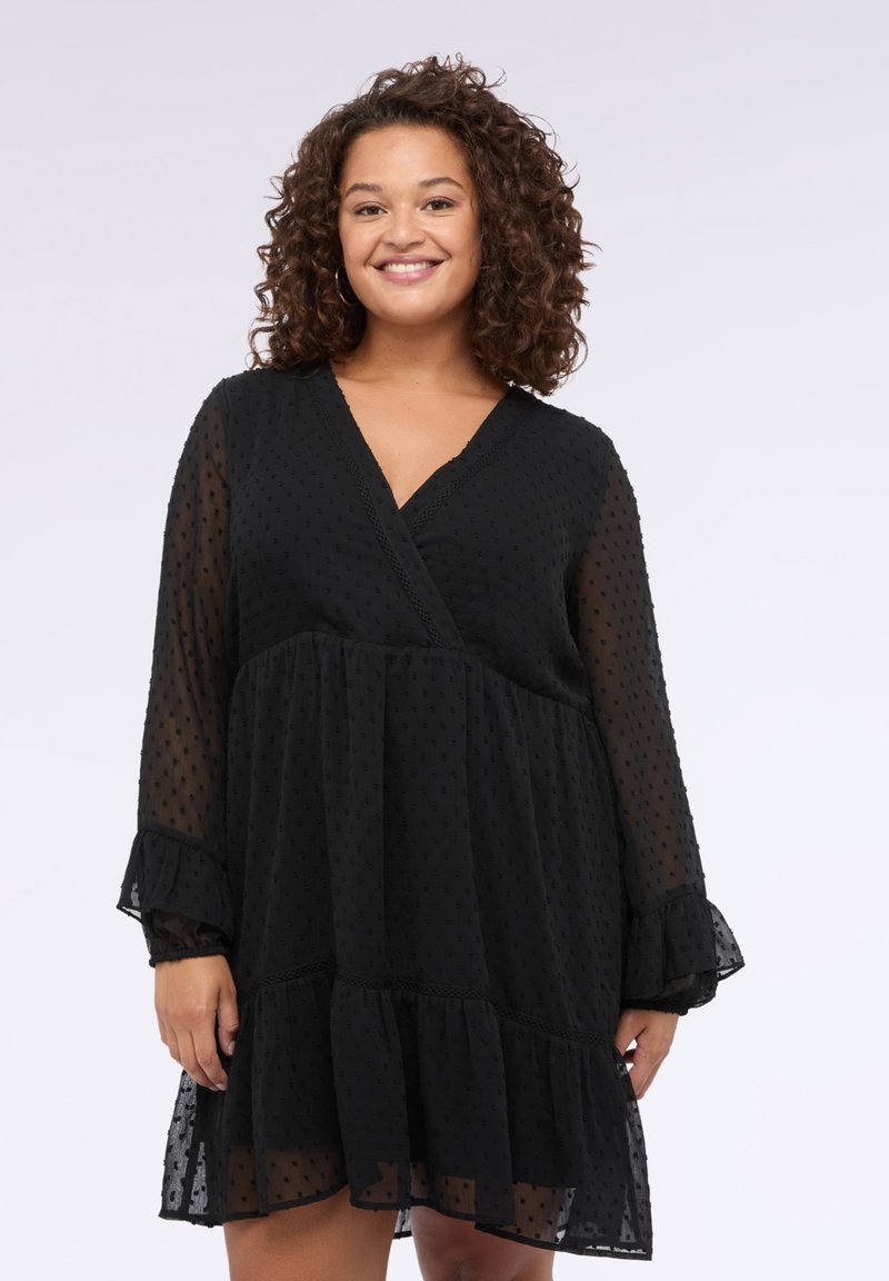 Vestido negro semitransparente con escote en V y mangas largas. Presenta pequeños puntos texturizados y un dobladillo con volantes. Tejido ligero con diseño en capas.