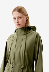 Χακί πράσινη αδιάβροχη ζακέτα με κουκούλα, ρυθμιζόμενα κορδόνια, κλείσιμο με κουμπί και πλαϊνές τσέπες. Λείος ματ υφή με ελάχιστο σχεδιασμό.