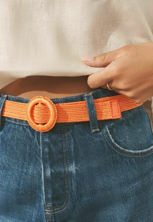 Hand adjusting beige top above blue jeans with an orange woven belt featuring a round buckle.