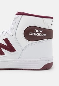 White high-top sneaker with dark burgundy accents. Smooth synthetic material, features prominent textured logo and robust lacing system.