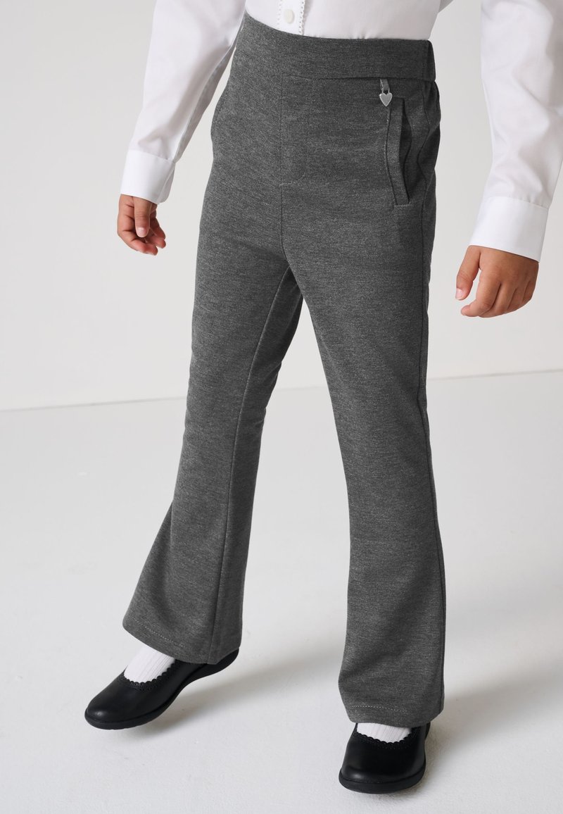 Kind trägt graue ausgestellte Hosen mit Seitentaschen, weißes Langarmshirt, weiße Socken und schwarze Schuhe und steht auf einem weißen Boden.