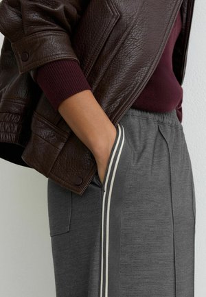 Hnědá kožená bunda přes vínové tričko, doplněná šedými tepláky s bílými pruhy na bocích a bočními kapsami.