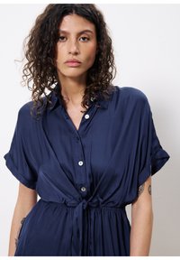 Navyblaues Hemdkleid mit kurzen, aufgekrempelten Ärmeln, Knoten-Detail in der Taille, seidig glatter Textur und subtiler glänzender Oberfläche.