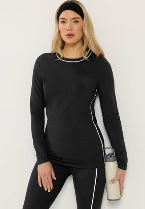 Blonde Frau in schwarzem Sportoutfit mit weißen Abnähern hält beige Wasserflasche, trägt schwarzen Stirnband und steht vor weißem Hintergrund.