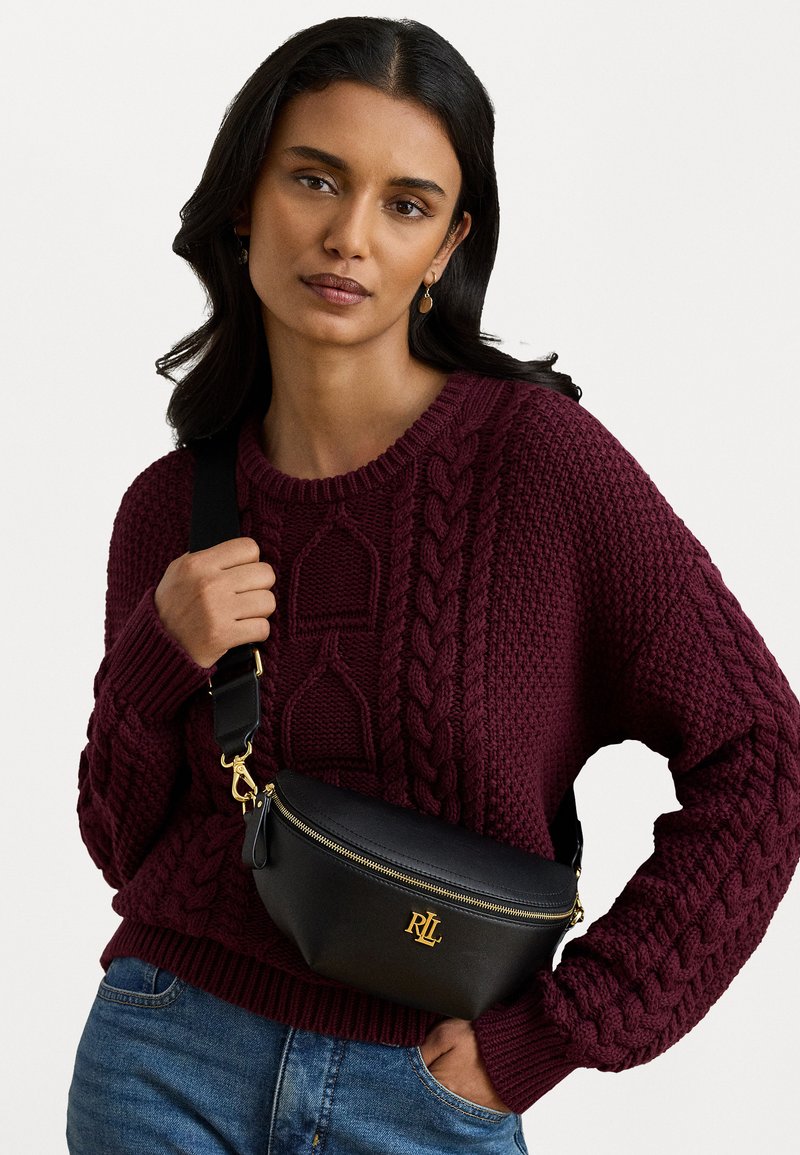 Μαύρη φτηνιασμένη δερμάτινη τσάντα μέσης με χρυσά αξεσουάρ, καμπυλωτό σχήμα και αντίθετη ραφή. Το μοντέλο φοράει πλεκτό πουλόβερ σε μπορντό απόχρωση.