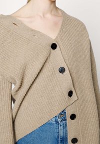 Cardigan beige en maille côtelée avec de gros boutons noirs, porté décalé sur une épaule, associé à un jean en denim bleu.