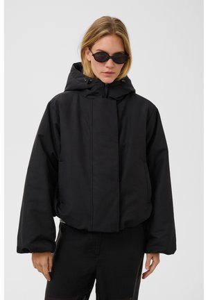 Kvinde iført en oversized sort hættetrøje og sorte solbriller, stående mod en ensfarvet grå baggrund.