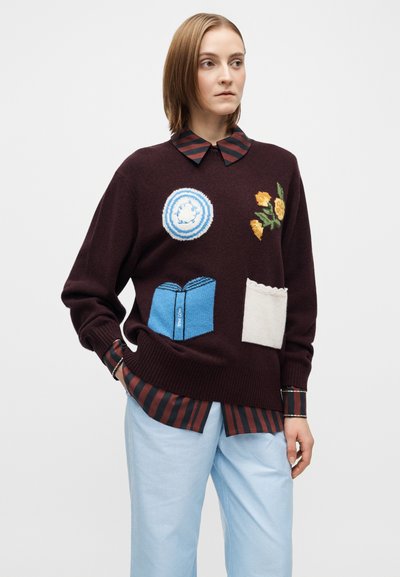 Vinrød strikket genser med mønstret broderi: et blått sirkulært motiv, gule blomster og en blå boklomme. Laget over en stripete skjorte.