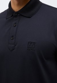 Polo noir avec trois boutons noirs et un logo brodé "66 North" ton sur ton sur le côté gauche de la poitrine.