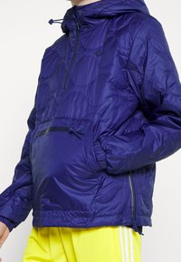 Lila puffjacka med huva, med halvdragkedja framtill, elastiska mudd och en ficka med dragkedja. Mönster med texturerade vågor och justerbar dragsko.