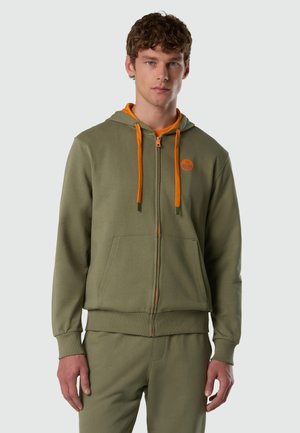 Giovane uomo che indossa una felpa con cappuccio verde oliva con cerniera e coulisse arancioni, accompagnata da pantaloni abbinati, rivolto verso la fotocamera su uno sfondo semplice.