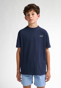 Camiseta de algodón azul marino de manga corta, con cuello redondo y un pequeño logo en el lado izquierdo del pecho; combinada con pantalones cortos azul claro.